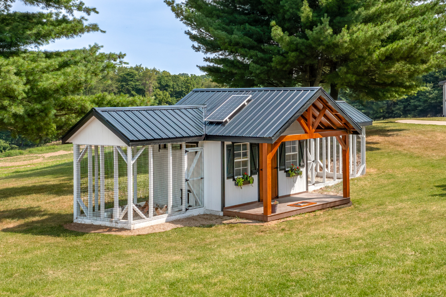 white combination coop with attached run on either side black metal roof with solar panel on it and a front porch leading to brown door and two windows with black shutters and flower boxes underneath